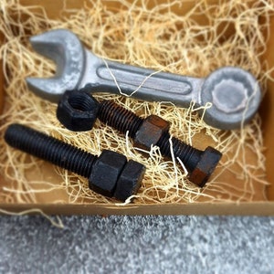 May include: A collection of metal tools, including a wrench, bolts, and nuts, presented in a wooden box filled with straw. The tools have a rustic, industrial appearance with a mix of silver and black finishes.