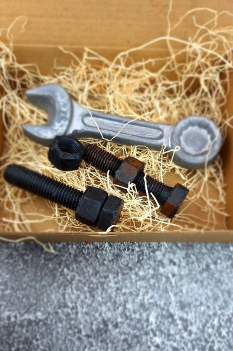 May include: A collection of metal tools, including a wrench, bolts, and nuts, presented in a wooden box filled with straw. The tools have a rustic, industrial appearance with a mix of silver and black finishes.