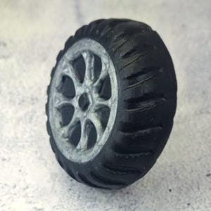 May include: A miniature black tire with a silver rim. The tire has a tread pattern and a detailed silver wheel design. The tire is a toy or model, likely for a vehicle.