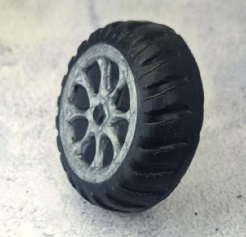 May include: A miniature black tire with a silver rim. The tire has a tread pattern and a detailed silver wheel design. The tire is a toy or model, likely for a vehicle.
