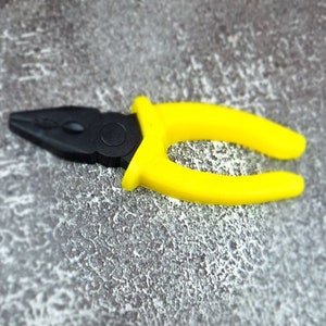 May include: A pair of pliers with a black head and bright yellow handles. The tool is designed for gripping and bending materials. The pliers are lying on a textured gray surface.