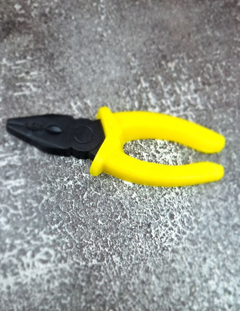 May include: A pair of pliers with a black head and bright yellow handles. The tool is designed for gripping and bending materials. The pliers are lying on a textured gray surface.