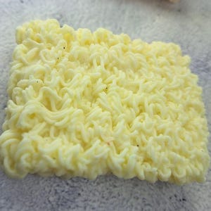 May include: Close-up of a square block of uncooked ramen noodles. The noodles are a pale yellow color and have a wavy, curly texture. Small dark specks are visible throughout the noodles.