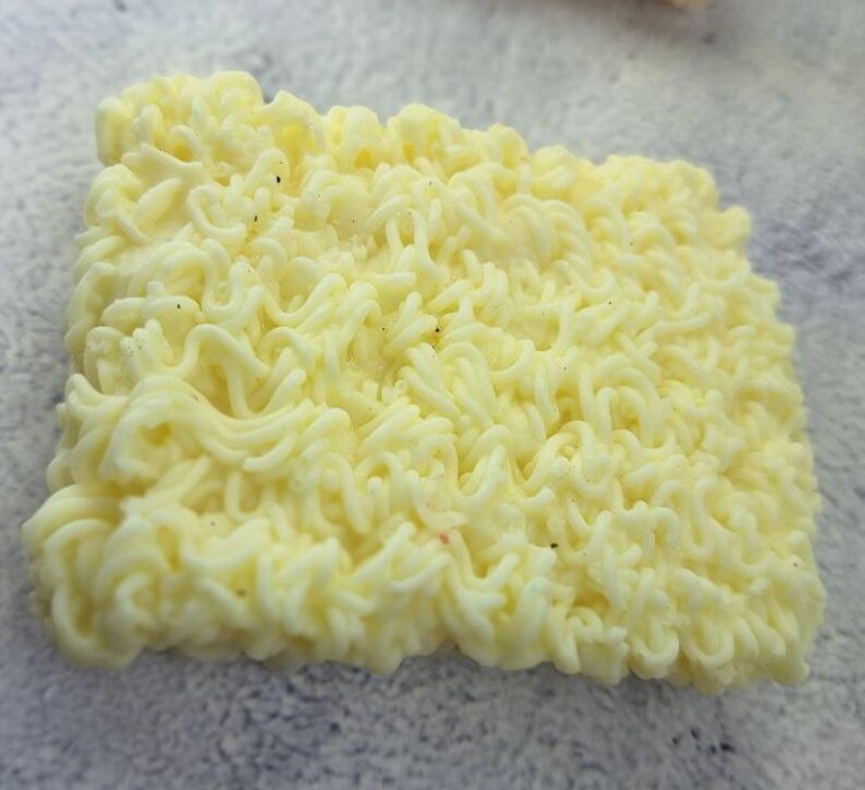 May include: Close-up of a square block of uncooked ramen noodles. The noodles are a pale yellow color and have a wavy, curly texture. Small dark specks are visible throughout the noodles.