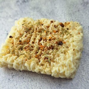 May include: A square block of uncooked ramen noodles, seasoned with herbs and spices. The noodles are a pale yellow color, and the seasoning includes red pepper flakes and dried herbs. The image is a close-up shot on a light gray surface.