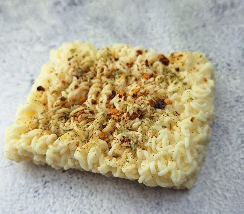 May include: A square block of uncooked ramen noodles, seasoned with herbs and spices. The noodles are a pale yellow color, and the seasoning includes red pepper flakes and dried herbs. The image is a close-up shot on a light gray surface.