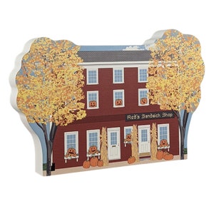 May include: A wooden model of a red brick building with a sign that reads "Red's Sandwich Shop". The building has windows with jack-o'-lantern faces and is surrounded by fall foliage and pumpkins.