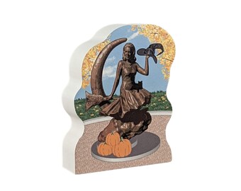 May include: A wooden figurine of a woman in a bronze-colored dress sitting on a crescent moon with a black cat and holding a witch's hat. The figurine is surrounded by orange pumpkins and set against a backdrop of fall foliage.