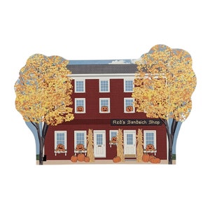 May include: A red brick building with a sign that reads "Red's Sandwich Shop". The building has windows with jack-o'-lantern faces and is decorated with pumpkins and corn stalks for Halloween.