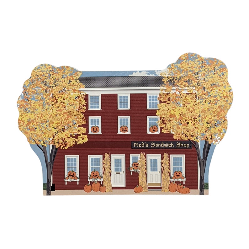 May include: A red brick building with a sign that reads "Red's Sandwich Shop". The building has windows with jack-o'-lantern faces and is decorated with pumpkins and corn stalks for Halloween.