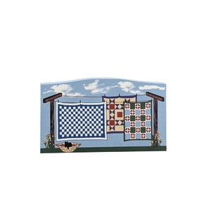 May include: A wooden wall art piece depicting a clothesline with three colourful quilts hanging on it. The quilts are a blue and white checked pattern, a red, yellow and green patchwork pattern, and a green and red checked pattern. The background is a blue sky with white clouds.
