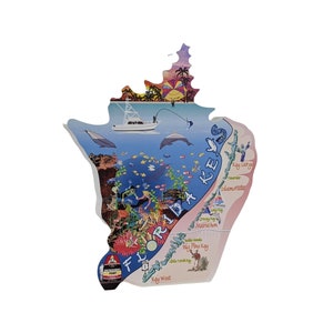 May include: A Florida Keys-shaped wooden wall art piece. The artwork depicts a vibrant underwater scene with fish, divers, and coral reefs. A boat, dolphins, and the text "Florida Keys" are also visible. The piece has a white border and a colorful sunset.