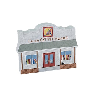 May include: A white building with a red brick base and a green roof. The building has a sign that reads "Calico Cat Patchworks". There is a black cat sitting in front of the building.