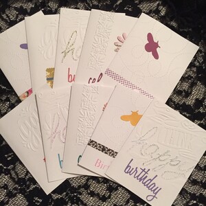 May include: A collection of white birthday cards with various designs. Some cards feature embossed floral patterns, while others have colorful butterfly cutouts or glittery text that says "Happy Birthday". The cards are arranged on a black lace surface.