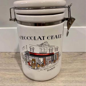 May include: White ceramic storage jar with a hinged lid and metal clasp. The jar is decorated with the words "Chocolat Chaud" and a colourful illustration of a Parisian cafe. The cafe scene includes people seated at tables.