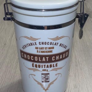 May include: A white ceramic canister with a lid, featuring the word "chocolat" and text in French. The canister has a metal clasp and a label that reads "Veritable Chocolat Belge Chocolat Chaud Equitable 400g."