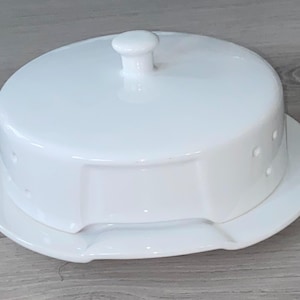 Op de afbeelding: Een witte keramische botervloot met een deksel en een kleine knop. De schaal staat op een bijpassend bord met een gebogen rand. Het ontwerp omvat kleine, gelijkmatig verdeelde gaten aan de zijkant van het deksel.