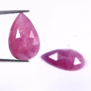May include: A pink pear-shaped gemstone with a faceted cut. The gemstone is held by tweezers and a reflection of the gemstone is visible below.