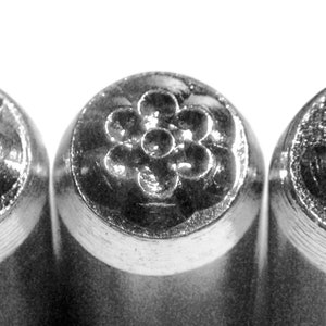 May include: Close-up of three metal stamps with floral and leaf designs. The stamps are cylindrical with detailed impressions of a cloud, a flower, and a leaf. The designs are raised and stand out against the metal.
