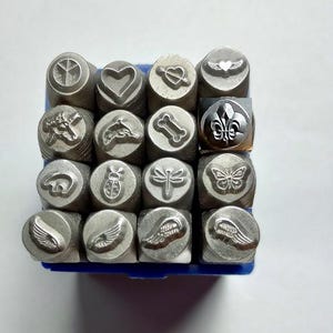May include: A collection of sixteen silver-toned metal stamping tools arranged in a four-by-four grid. Each tool features a unique embossed design on its circular face, including a peace sign, heart, arrow through heart, winged heart, unicorn, dolphin, bone, fleur-de-lis, bear, ladybug, dragonfly, butterfly, and four distinct wing patterns.