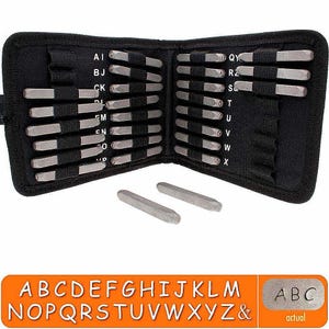 May include: A black fabric case, open to display a set of metal letter stamps. The stamps are arranged in rows and held in place by elastic bands. Two loose stamps are at the bottom. An orange rectangle displays the alphabet and the letters "ABC" stamped.