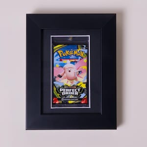 May include: A black rectangular frame containing a sealed Pokémon trading card booster pack. The pack features a pink character, yellow accents, and the text Pokémon Trading Card Game and Mega Evolution Perfect Order on a dark background.