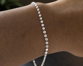 Könnte beinhalten: Ein zartes Tennisarmband mit einer durchgehenden Reihe kleiner, runder, klar facettierter Steine, die in eine dünne, polierte goldfarbene Metallkette gefasst sind und das Handgelenk umschließen, was für einen dezenten Look mit funkelnden Lichtreflexen sorgt.