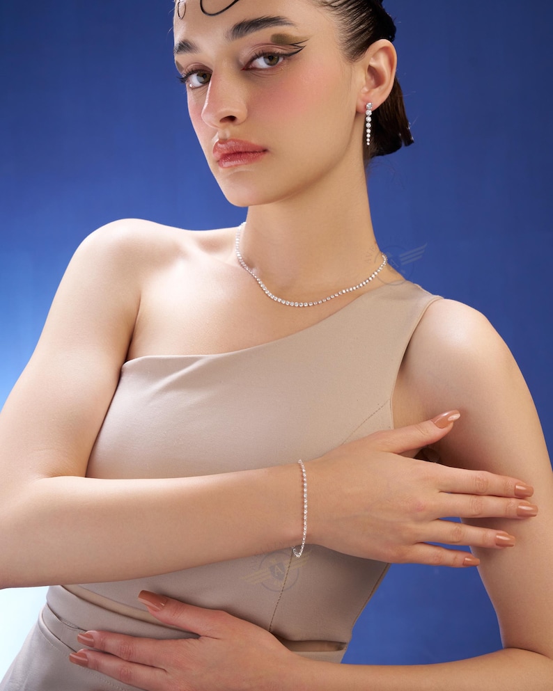 K&ouml;nnte beinhalten: Ein Schmuckset bestehend aus einer silberfarbenen Tennis-Halskette, einem passenden Tennis-Armband und einem Paar h&auml;ngender Ohrringe. Jedes St&uuml;ck zeigt eine durchgehende Reihe kleiner, klarer, rund geschliffener Steine in einer Metallfassung.