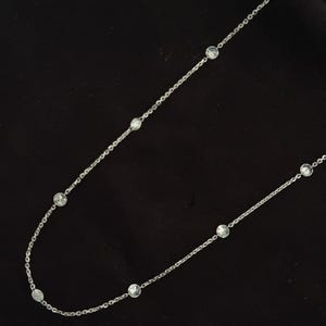 K&ouml;nnte beinhalten: Eine zarte silberfarbene Kettenhalskette mit gleichm&auml;&szlig;ig verteilten, runden, klaren facettierten Steinen, die in Metallfassungen entlang der Kette eingesetzt sind. Die Halskette wird vor einem dunklen, einfarbigen Hintergrund pr&auml;sentiert.