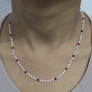 Op de afbeelding: Een delicate ketting met afwisselende heldere en rode edelstenen. De ketting bestaat uit kleine, ronde, heldere stenen afgewisseld met levendige rode stenen. De ketting wordt om de nek gedragen en toont het elegante ontwerp en het kleurcontrast.