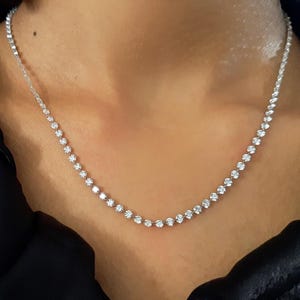 May include: A silver-toned tennis necklace featuring a continuous row of small, round, clear, faceted stones set in a metallic chain. The necklace rests against the neck, displaying a uniform arrangement of sparkling stones along the entire length.