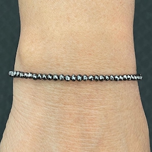 May include: A delicate bracelet composed of small, faceted, dark gray beads. The beads are strung together to form a single strand, creating a minimalist and elegant accessory. The bracelet is shown on a wrist.