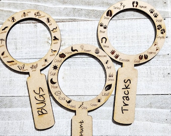 May include: Three wooden magnifying glass-shaped scavenger hunt tools with different themes: Bugs, Tracks, and Scavenger Hunt. Each tool has a circle with various images and text labels around the edge.