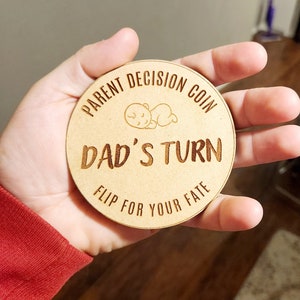 May include: A wooden coin with the text "Parent Decision Coin" and "Dad's Turn" with a small illustration of a sleeping baby. The text "Flip for your fate" is also on the coin.