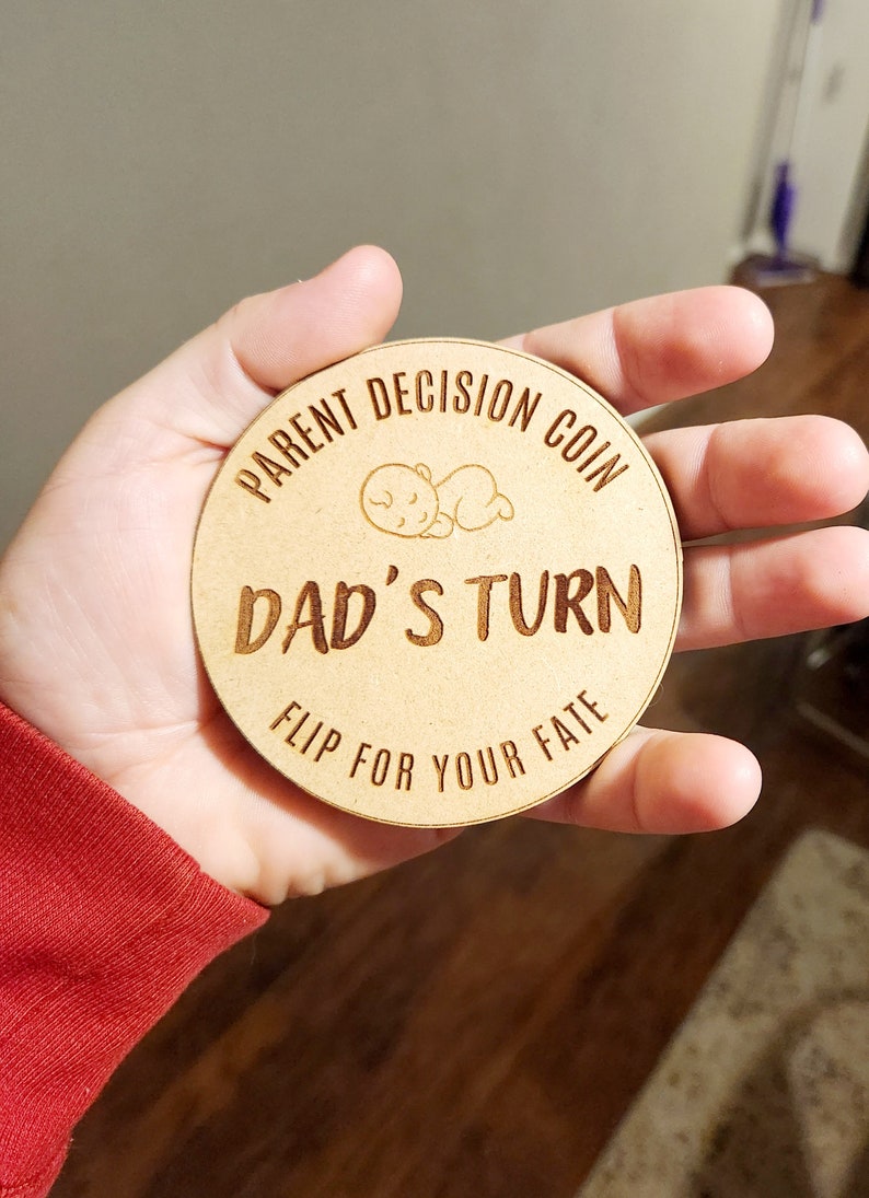 May include: A wooden coin with the text "Parent Decision Coin" and "Dad's Turn" with a small illustration of a sleeping baby. The text "Flip for your fate" is also on the coin.