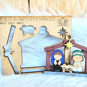 Puede incluir: Kit de manualidades de madera para construir un pesebre de Navidad. El kit incluye un pesebre, figuras de María, José y una oveja, y una estrella. El kit está pintado en marrón, azul y verde. El texto "BUILD-A-NATIVITY MANGER" está impreso en la madera. El texto "PAINT IT - GLUE IT - HANG IT" está impreso en la madera. El texto "Oh Holy Night" está impreso en la madera.