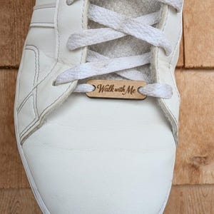 May include: Close-up of a white sneaker with white laces and a wooden tag that reads "Walk with Me". The shoe is on a wooden surface.