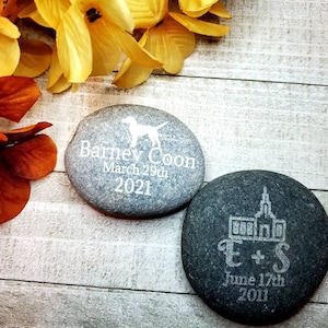 May include: Two engraved river rocks. One gray rock with a dog silhouette and the text "Barney Coon March 29th 2021". One black rock with a church silhouette and the text "E+S June 17th 2011".
