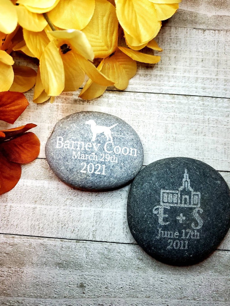 May include: Two engraved river rocks. One gray rock with a dog silhouette and the text "Barney Coon March 29th 2021". One black rock with a church silhouette and the text "E+S June 17th 2011".
