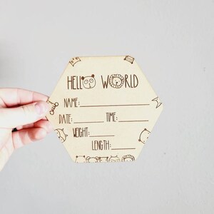 Könnte beinhalten: Ein sechseckiges Schild aus Holz mit dem Text "Hello World" und Platz für den Namen, das Datum, die Uhrzeit, das Gewicht und die Länge des Babys. Das Schild ist mit Comic-Tieren verziert.