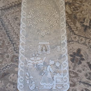May include: A white lace kitchen curtain with the words "MOTHER'S KITCHEN" in a circular design at the top. The curtain features a detailed scene of a woman cooking, a table, and a cat. The edges are scalloped.