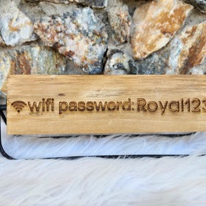 May include: A wooden sign displaying the text "wifi password: Royal123". The sign is made of light-coloured wood and is supported by a black metal frame. The background features a stone wall.