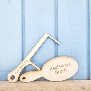 Könnte beinhalten: Ein Holzkamm- und -bürstenset mit dem eingravierten Text "Bald Man's Comb" und "Bald Man's Brush". Der Kamm und die Bürste liegen auf einer Holzoberfläche vor einer hellblauen Wand.
