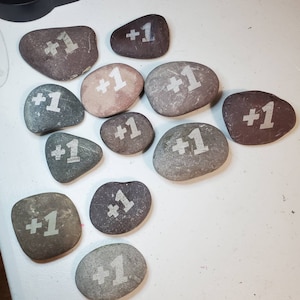 May include: A collection of 10 smooth, gray and brown stones with the words " +1 " painted in white on each stone.