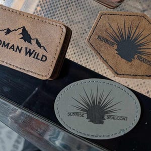May include: Three rectangular, pentagonal, and oval-shaped patches with stitched edges. One tan rectangular patch features a mountain graphic and the text ROMAN WILD. Two tan and gray patches feature a sunburst graphic and the text SUNRISE SEALCOAT.
