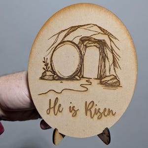 Könnte beinhalten: Runde Osterdekoration aus Holz mit einem lasergravierten Design eines leeren Grabes und dem Schriftzug "He is Risen". Die Dekoration wird von einem kleinen Holzständer getragen. Das Design zeigt einen vom Grab weggerollten Stein.