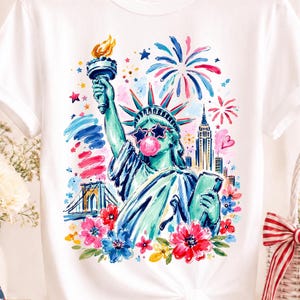 Op de afbeelding: Wit T-shirt met een kleurrijke illustratie van het Vrijheidsbeeld met een sterrenbril en een kauwgomballenbel. Vuurwerk, bloemen en skyline maken het ontwerp compleet. Het shirt hangt aan een houten hanger.
