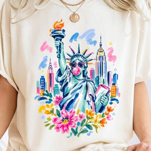 Könnte beinhalten: Cremefarbenes T-Shirt mit einer farbenfrohen Illustration der Freiheitsstatue mit Sonnenbrille und einer Kaugummiblase. Das Kunstwerk umfasst eine Stadtlandschaft und florale Akzente, die ein lebendiges und verspieltes Design schaffen.