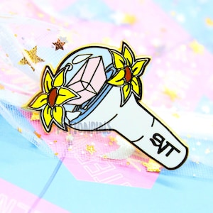 Può includere: Uno spillo in smalto blu e bianco di un bastone luminoso con girasoli gialli. Il bastone luminoso ha il testo "SVT" su di esso.