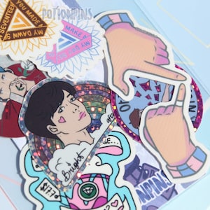 Può includere: Una collezione di adesivi colorati con la band K-pop Seventeen. Gli adesivi includono un adesivo olografico arcobaleno con il testo "Bright", un adesivo con un design a girasole e il testo "You Made My Dawn", un adesivo con una mano che fa un cuore, e un adesivo con il testo "I Know I Can't Take It".